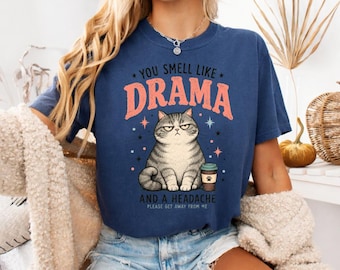 May include: A navy blue short-sleeve t-shirt featuring a graphic of a grumpy grey tabby cat holding a coffee cup. The text reads YOU SMELL LIKE DRAMA AND A HEADACHE PLEASE GET AWAY FROM ME. The design includes small star accents in pink and blue.