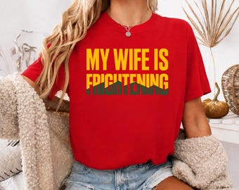 May include: A red short-sleeved t-shirt featuring the yellow text "MY WIFE IS" above the green text "FRIGHTENING" in a bold, sans-serif font. The shirt has a crew neckline and is worn with light blue denim shorts.