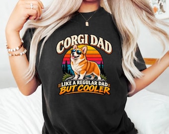 May include: A black t-shirt featuring a graphic of a Corgi wearing sunglasses. The design includes a sunset background with silhouettes of trees and the text "CORGI DAD" at the top and "LIKE A REGULAR DAD BUT COOLER" at the bottom in bold letters.