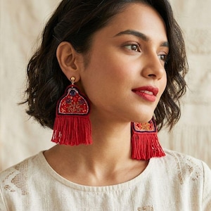 Puede incluir: Un par de pendientes llamativos con una parte superior de tela bordada en rojo con un patrón floral y una piedra central de color púrpura oscuro, terminados con una borla de flecos rojos largos y densos, sujetos con un perno dorado.