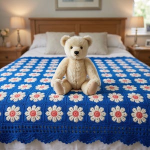 Puede incluir: Un oso de peluche de color crema sentado sobre una manta de ganchillo hecha a mano que presenta un patrón repetitivo de margaritas blancas y color coral sobre un fondo azul vibrante, con un borde festoneado decorativo en la parte inferior.