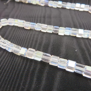 May include: Strands of iridescent, rectangular beads. The beads are clear with a subtle rainbow sheen, strung together to form a flexible chain. The background is a dark, textured fabric.