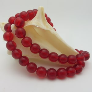 May include: Two strands of red beaded jewelry are draped over a large, off-white seashell. The beads are round and have a translucent quality, with a deep red color. The seashell is smooth and has a natural, organic shape.