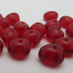 May include: A group of red glass beads, each with a small hole in the center. The beads are scattered on a white surface.
