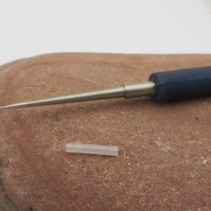 May include: A metal awl with a black handle and a sharp, pointed tip. A small, translucent plastic tube is next to the awl. The objects are on a reddish-brown, textured surface.