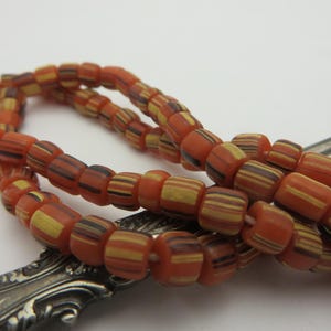 May include: Close-up of a necklace with orange, yellow, and brown striped beads. The beads are cylindrical and strung together. The necklace is resting on a silver object, possibly a spoon or fork. The beads have a vintage or antique appearance.