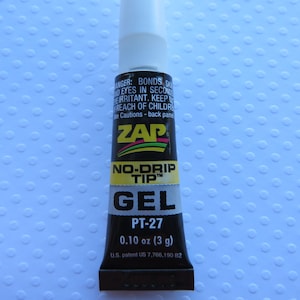 May include: A black tube of ZAP No-Drip Tip Gel glue with a yellow label. The label says "PT-27" and "0.10 oz (3 g)". The tube is on a white background with a pattern of small circles.