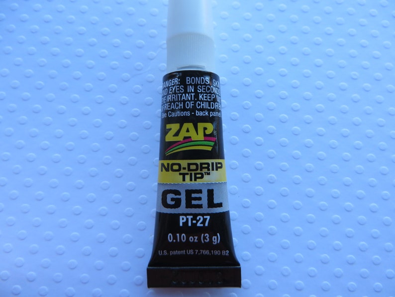 May include: A black tube of ZAP No-Drip Tip Gel glue with a yellow label. The label says "PT-27" and "0.10 oz (3 g)". The tube is on a white background with a pattern of small circles.