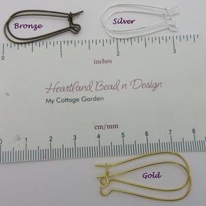 May include: Three earring wires, one bronze, one silver, and one gold, are shown against a white background with a ruler for size reference. The ruler is marked in inches and centimeters.