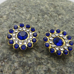 May include: Two gold-toned, flower-shaped embellishments with a central large blue gem surrounded by smaller white and blue gems. The embellishments are set against a gray stone background.