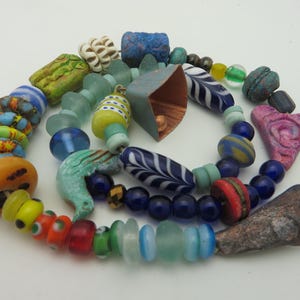 May include: A collection of colorful, handcrafted beads in various shapes and sizes. The beads include shades of blue, green, yellow, red, and orange. Some beads have unique patterns and textures, including stripes, swirls, and leaf-like designs. A copper-colored element is also present.