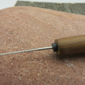 May include: A metal needle tool with a thin, serrated shaft and a dark brown wooden handle. The tool features a small black circular collar where the metal shaft meets the wood. The handle is cylindrical and tapers slightly toward the metal tip.