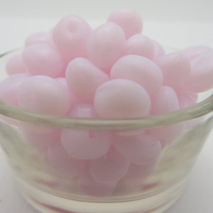 May include: A clear glass bowl filled with light pink, oval-shaped beads. The beads have a smooth, glossy texture and are clustered together. The bowl is set against a plain white background, highlighting the beads' color and shape.
