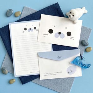 May include: A stationery set featuring a white lined sheet and two envelopes decorated with a cartoon seal face. The envelopes include the text "A fluff of snow on rock's cold palmwhiskers catch" and a small paw print icon on the flap.