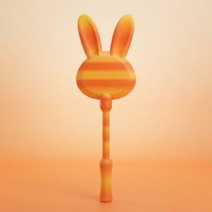 May include: A handheld wand featuring a rounded head with two upright rabbit ears, decorated with horizontal orange and yellow stripes. The slender handle also displays matching orange and yellow stripes, ending in a smooth, solid orange base.