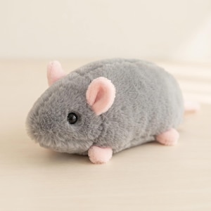 Könnte beinhalten: Ein Plüschtier in Form einer kleinen Maus mit weichem grauem Fell, abgerundeten hellrosa Ohren und kleinen rosa Füßen. Die Maus hat ein sichtbares schwarzes Auge und einen abgerundeten Körper aus flauschigem Material mit kompaktem Design.