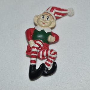 May include: A decorative elf figurine featuring a red and white striped hat with a pom-pom, a green vest, a white collar, and red sleeves. The elf has red and white striped legs and wears black pointed shoes, with a smiling face and large ears.