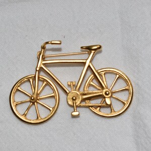 May include: A gold-toned metal bicycle brooch featuring a detailed frame, two spoked wheels, handlebars, a seat, and a pedal mechanism. The pin closure is visible along the top bar of the bicycle frame, providing a secure way to attach the accessory.