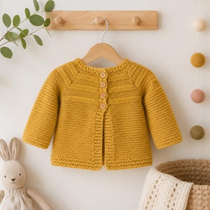 May include: A mustard yellow long-sleeve knit cardigan featuring a round neckline and a vertical row of four small wooden buttons down the centre front. The garment is constructed with a textured garter stitch pattern and hangs on a wooden hanger.