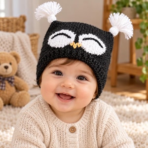 May include: A black knitted beanie featuring a white owl face design with closed eyes, a small yellow stitched beak, and two white yarn tassels attached to the top corners, resembling owl ears. The hat is crafted with a textured, chunky knit pattern.
