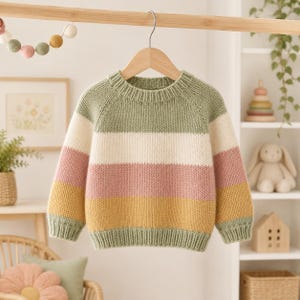 May include: A long-sleeved knit jumper featuring horizontal stripes in sage green, cream, dusty rose, and mustard yellow. The garment has a ribbed crew neckline, ribbed cuffs, and a ribbed hem, all displayed on a wooden hanger.