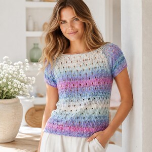 May include: A short-sleeved knit top featuring a variegated color pattern of soft blue, purple, and white hues. The garment is constructed with a delicate open-stitch texture, a rounded neckline, and raglan sleeves, creating a lightweight appearance.