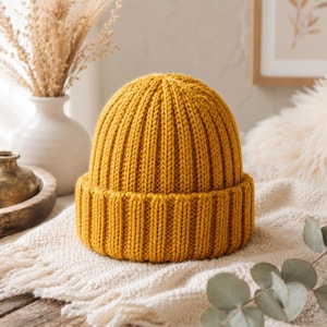 Könnte beinhalten: Eine senfgelbe Beanie-Mütze mit einem dicken, vertikalen Rippstrickmuster und einem breiten, umgeschlagenen Bündchen an der Basis. Die Mütze ist aus einer gleichmäßigen, groben Garnstruktur gefertigt, die im Ruhezustand eine strukturierte, runde Form bildet.