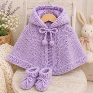 May include: A light purple hand-knitted hooded cape featuring a garter stitch border and a central drawstring closure with two pom-poms, accompanied by a matching pair of knitted booties with small decorative bows on the front of each shoe.