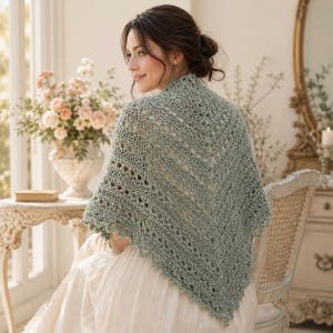 May include: A sage green triangular shawl featuring an intricate openwork crochet pattern with a scalloped edge. The garment is draped over the shoulders, showcasing a textured, lightweight knit construction with a pointed hemline at the back.