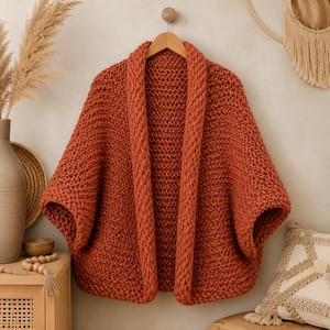 Puede incluir: Un cárdigan de punto grueso en un color naranja quemado sólido. Esta prenda de frente abierto presenta un cuello chal grueso y acanalado, junto con mangas anchas y holgadas. La textura está creada con hilo pesado, formando un patrón de punto uniforme.