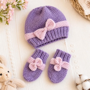 May include: A knitted purple beanie hat and a pair of matching mittens. The beanie features a light pink knitted bow centered on a textured band. Each mitten is adorned with a matching light pink knitted flower detail on the cuff area.