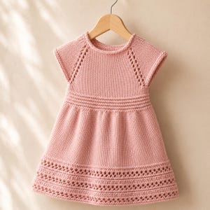 Può includere: Un abito lavorato a maglia color rosa polvere con maniche corte, scollo rotondo e corpetto testurizzato. Il capo presenta motivi decorativi traforati lungo le maniche raglan e un ampio e intricato bordo traforato sull'orlo inferiore.