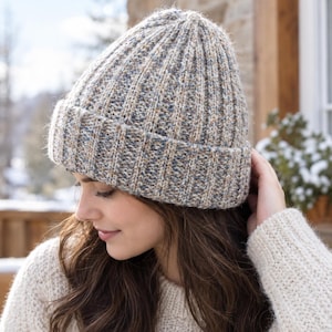 Puede incluir: Un gorro de punto acanalado en una mezcla jaspeada de hilos beige, gris y azul. El gorro presenta un puño grueso doblado en la base y una textura acanalada vertical en toda la superficie, confeccionado con hilo multicolor de aspecto moteado.