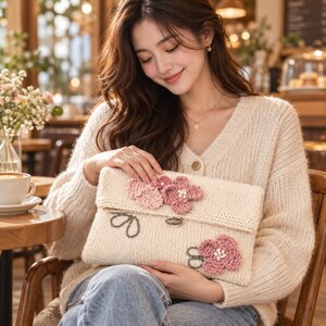 Puede incluir: Un bolso de mano de punto color crema con una solapa plegable adornada con flores de ganchillo tridimensionales en tonos rosa y malva, acentuado con tallos verdes bordados y un cierre central de metal ovalado.