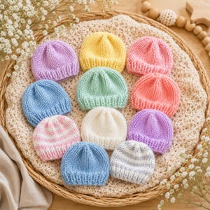 May include: A collection of twelve small, hand-knitted beanies in various solid colors including yellow, lavender, light blue, mint green, pink, and white, along with two striped patterns, arranged on a textured, cream-colored fabric surface.
