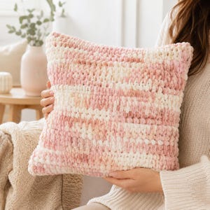 May include: A square throw pillow made of thick, plush yarn in a variegated pattern of soft pink, cream, and white. The pillow features a chunky, hand-knitted texture with visible loops throughout, creating a soft and tactile surface appearance.