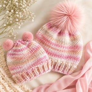 May include: Two knitted beanies featuring a variegated yarn pattern in shades of pink, white, and tan. The larger beanie has a single large pink faux fur pom-pom on top, while the smaller beanie features two smaller matching pink pom-poms.