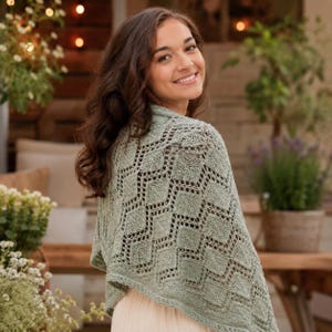 May include: A sage green knitted shawl featuring a repeating geometric diamond pattern with openwork lace detailing. The garment is draped over the shoulders, showcasing a textured knit construction with a consistent light green colour throughout.