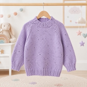Könnte beinhalten: Ein langärmeliger, hellvioletter Strickpullover mit geripptem Stehkragen, gerippten Bündchen und einem gerippten Saum. Der Pullover ist aus einem Garn mit gesprenkelter Textur gefertigt und weist eine klassische Raglan-Ärmelkonstruktion auf.