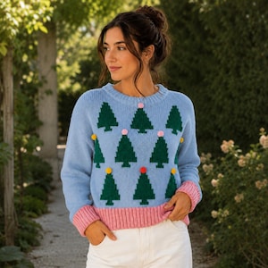 May include: A light blue long-sleeve sweater featuring a repeating pattern of dark green pixelated Christmas trees. The garment includes a ribbed pink hem and cuffs, and is decorated with small, colorful pom-poms scattered across the front.