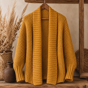 May include: A mustard yellow open-front cardigan featuring a chunky knit texture, a wide ribbed shawl collar, and long sleeves with fitted ribbed cuffs. The garment is displayed on a wooden hanger, showcasing a consistent knit pattern throughout.