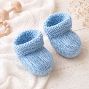 Pode incluir: Um par de sapatinhos de bebé em malha azul-clara. Os sapatinhos apresentam um design clássico de biqueira arredondada com uma dobra no tornozelo, exibindo uma textura consistente de ponto musgo em toda a estrutura do calçado.