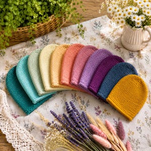 May include: A collection of ten knitted beanies arranged in a fan shape, featuring a spectrum of colours including teal, light green, cream, yellow, peach, pink, lavender, purple, navy blue, and mustard yellow, all made with a simple knit stitch.