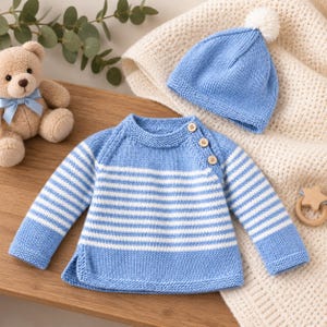 Puede incluir: Un suéter de punto de manga larga a rayas azul claro y blanco con tres pequeños botones de madera en el hombro, junto a un gorro de punto azul claro a juego con un pompón blanco, presentado junto a un oso de peluche.