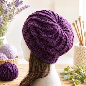 May include: A deep purple knitted beanie featuring a textured spiral pattern on the crown and a ribbed brim. The hat is displayed on a mannequin head, showcasing the intricate stitch work and the rich, saturated color of the yarn used throughout.
