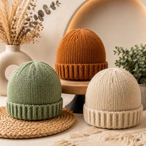 Puede incluir: Tres gorros de punto en colores sólidos: uno verde salvia, uno naranja quemado y uno crema. Cada gorro presenta un patrón de punto acanalado vertical en la corona y un puño acanalado grueso y doblado en la base, ofreciendo una apariencia texturizada.