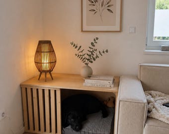 Puede incluir: Una caseta para mascotas de madera clara con diseño de listones y superficie superior plana. Un perro negro descansa dentro sobre un cojín gris. Encima hay una lámpara de bambú marrón, un jarrón beige con ramas verdes y dos libros.