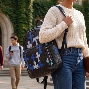 Op de afbeelding: Een zwarte rugzak met een herhalend patroon van blauwe en witte kruisvormige patches, gedragen met een crèmekleurige kabeltrui, een blauwe spijkerbroek en een bruin boek in de hand, voor een stenen universiteitsgebouw.