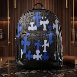 Puede incluir: Una mochila de cuero negro con un patrón de parches superpuestos en forma de cruz en tonos azul oscuro, azul claro y negro. La bolsa incluye un asa superior, correas ajustables para los hombros y un bolsillo frontal con cremallera y tirador azul.
