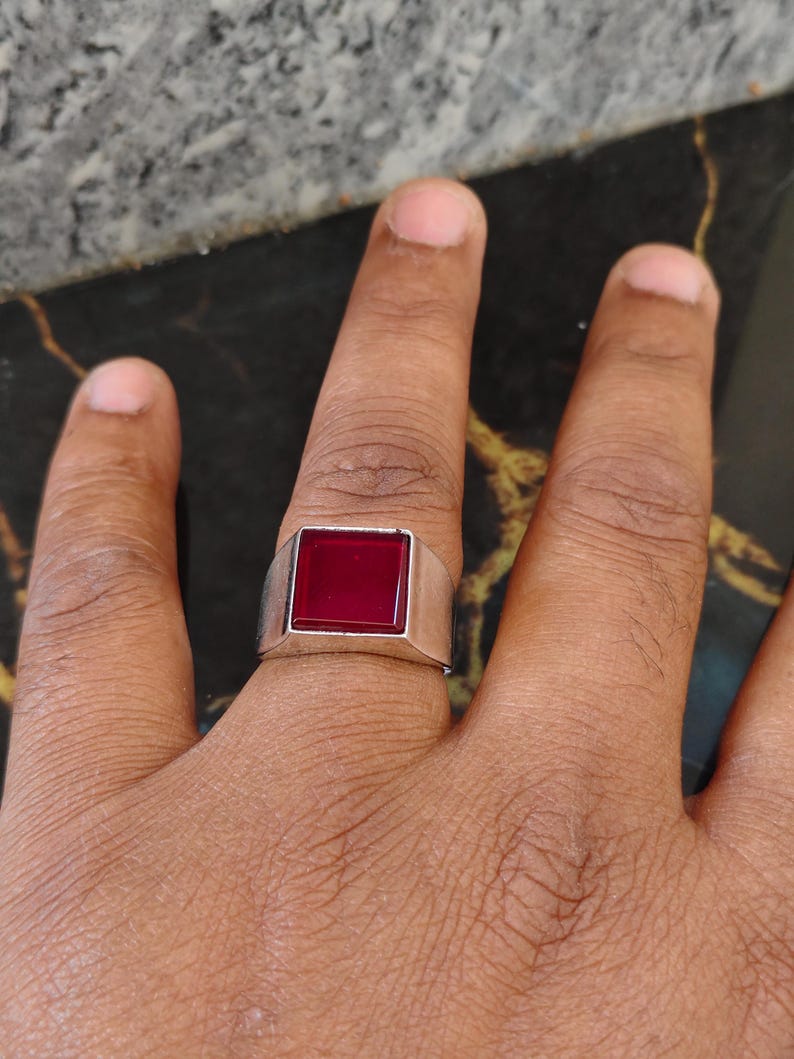 May include: A silver-toned metal ring featuring a prominent, square-shaped, deep red gemstone set in the center. The band of the ring is smooth and solid, with a wide, flat-topped design that holds the square red stone securely in place.